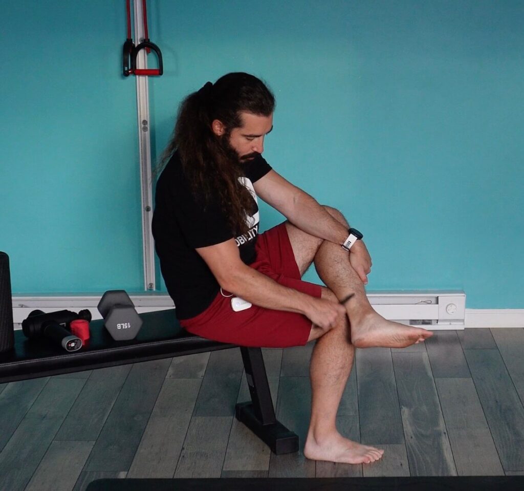 Dr. John is seated with his left leg over his right in a figure 4. He is using muscle scraping in the achilles tendon to promote healing and blood flow for achilles tendinitis self-care